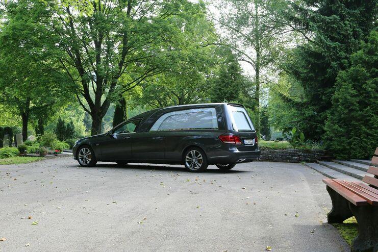 Dunkler Leichenwagen parkt auf einem Weg zwischen Bäumen auf einem Friedhof, neben einer Bank.