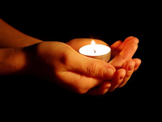 Hände halten eine flackernde Teelichtkerze vor einem dunklen Hintergrund.