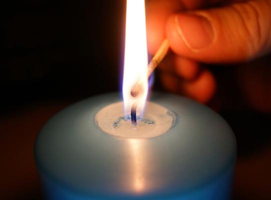 Nahaufnahme einer brennenden Kerze mit einer Hand, die ein Streichholz zur Flamme hält.