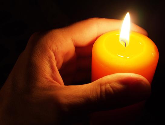 Eine Hand hält eine brennende orangefarbene Kerze in einem dunklen Umfeld.