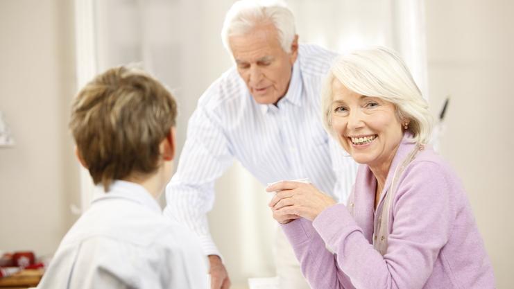 Lächelnde ältere Frau sitzt am Tisch, während ein Mann im Hintergrund Getränke serviert, ein Junge schaut sie an.